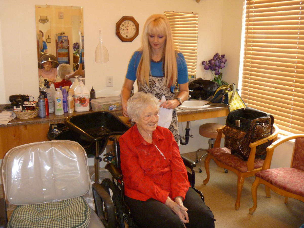 An elderly woman in a wheelchair smiles while getting her hair styled at a salon.