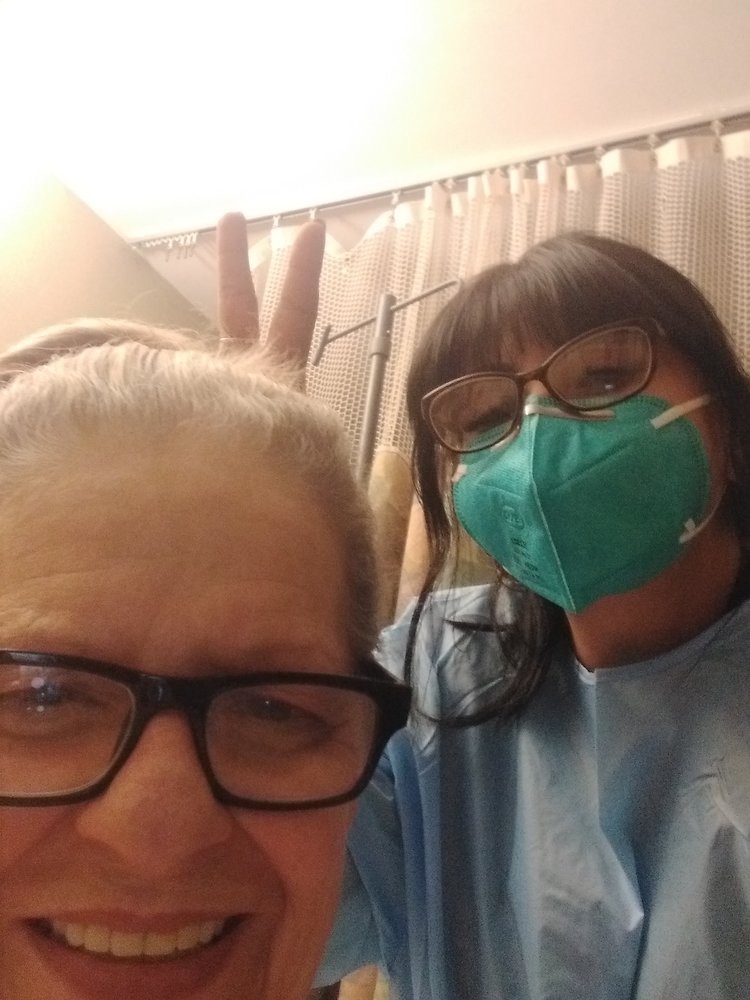A person taking a selfie with another individual in a medical setting, both wearing glasses and one wearing a mask.