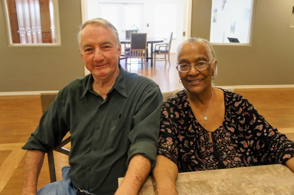 An elderly couple sitting together indoors and smiling at the camera.
