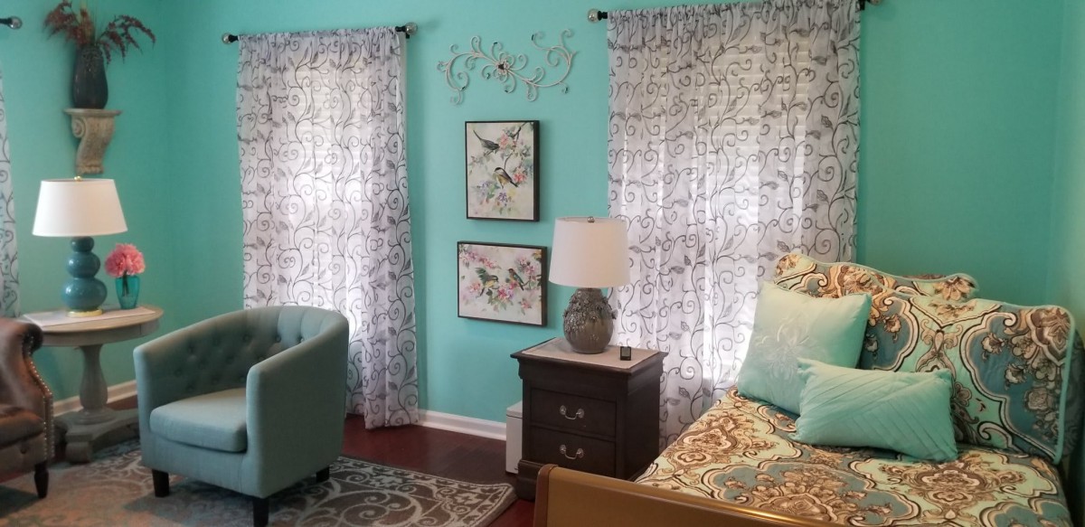 A cozy bedroom featuring turquoise walls, floral curtains and bedding, a nightstand with a lamp, and decorative wall art.