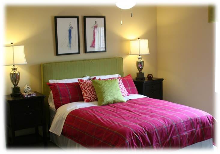 A cozy bedroom with a green headboard, red bedding, and colorful pillows, featuring two framed artworks above the bed and a lamp on the bedside table.