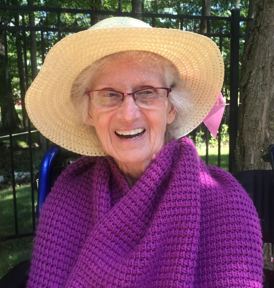 An elderly woman smiling while wearing a large straw hat and a purple shawl, sitting outdoors.