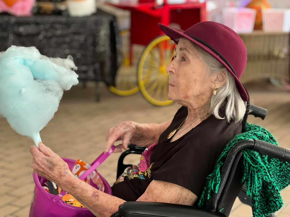 An elderly woman in a wheelchair enjoys cotton candy indoors, wearing a stylish hat.