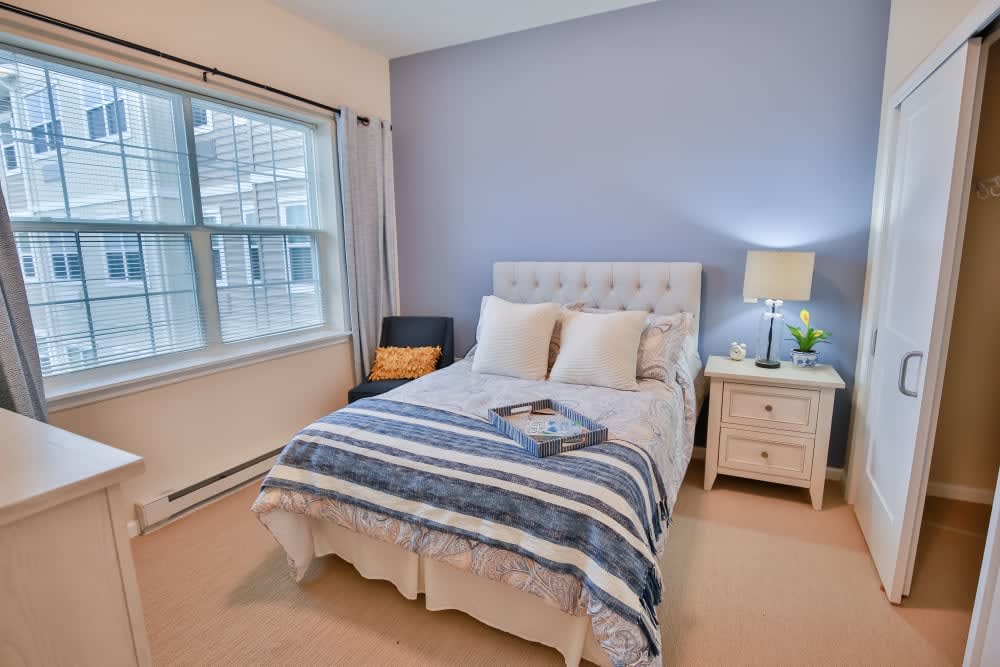 A cozy bedroom featuring a neatly made bed with striped bedding, a nightstand with a lamp, and a window letting in natural light.