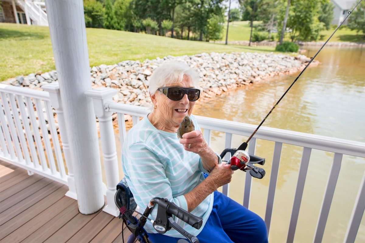 A person sitting in a wheelchair is fishing by a lake, wearing sunglasses and holding a fish.