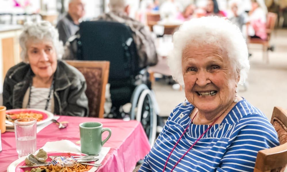An elderly woman smiling at a dining table in a nursing home.