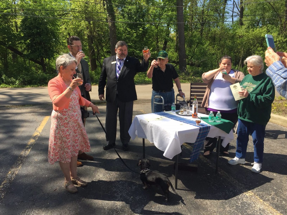 A group of people gathered outdoors at an event, with a woman speaking into a microphone and a dog nearby.