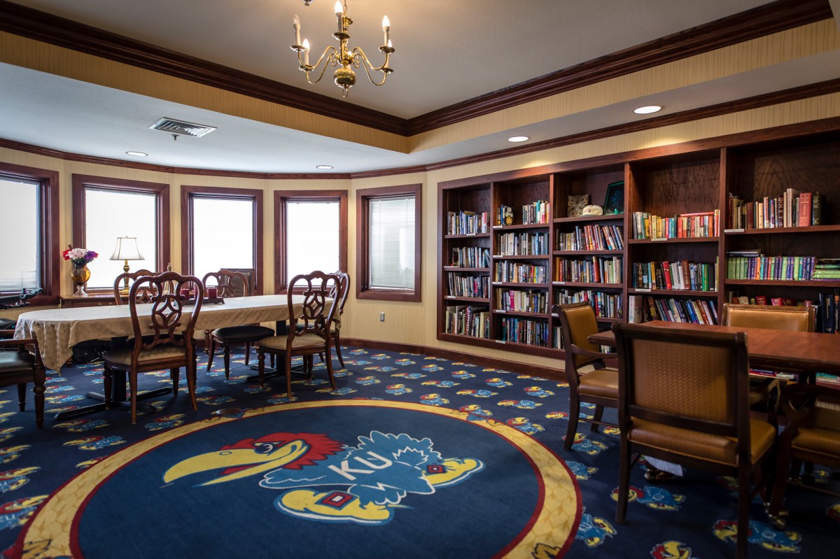 A cozy library room with bookshelves, a table, chairs, and a colorful carpet featuring a bird logo.