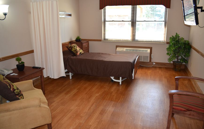 A hospital room featuring a single bed, wooden flooring, and a window with blinds.