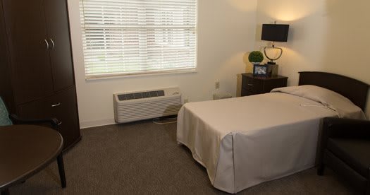 A cozy bedroom with a bed, side table, lamp, and an air conditioner beneath the window.