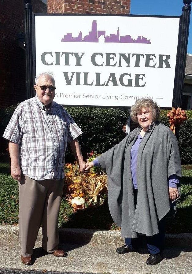 An elderly couple stands in front of a 'City Center Village' sign outdoors.