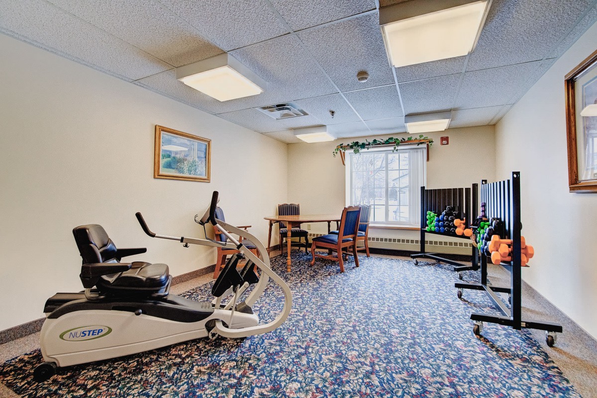 A small exercise room with a stationary bike, weights, and chairs on a carpeted floor.