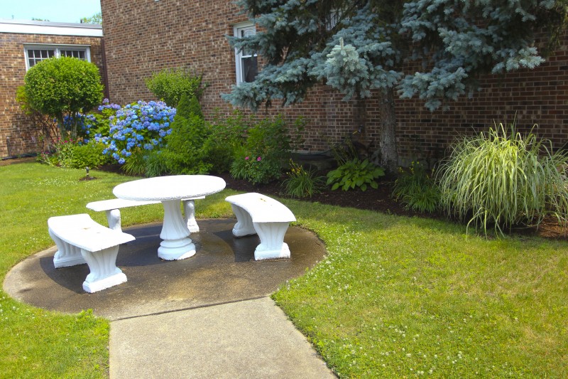 A peaceful garden setting featuring a white stone table with benches surrounded by lush greenery and bushes.