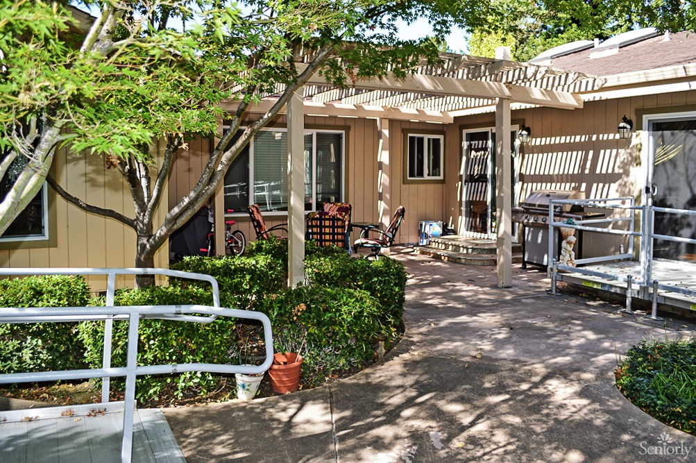A cozy outdoor patio area with a pergola, surrounded by trees and plants, featuring seating arrangements and dappled sunlight.