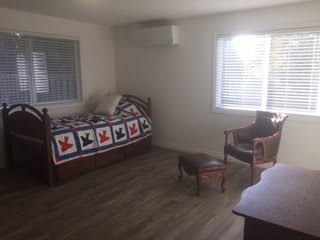 A minimalist bedroom with a daybed featuring a colorful quilt, a wooden chair, and natural light streaming through two windows.