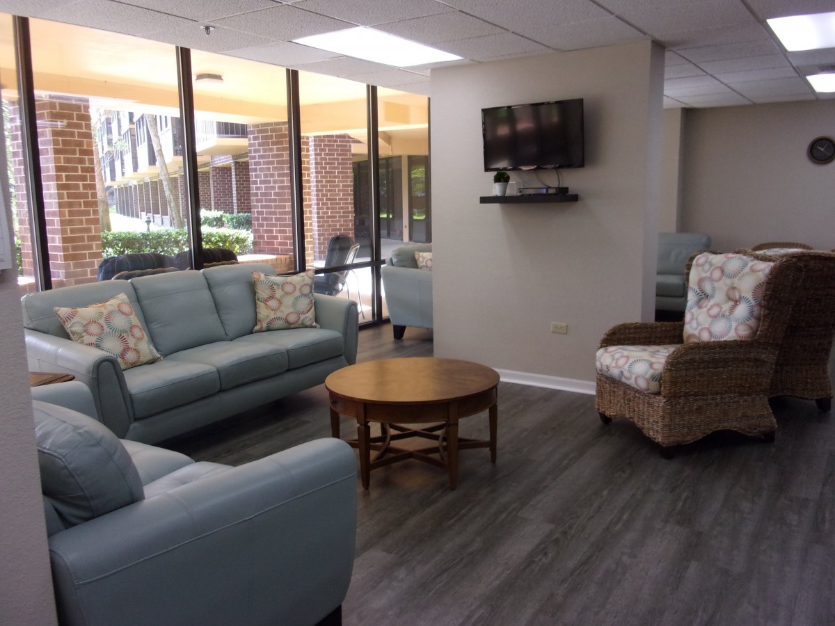 A modern living room with sofas, a coffee table, and a wall-mounted TV, featuring large windows that let in natural light.