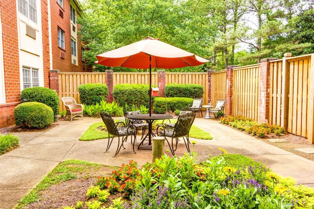 A cozy patio area with a table and chairs under a large umbrella, surrounded by lush greenery and a wooden fence.