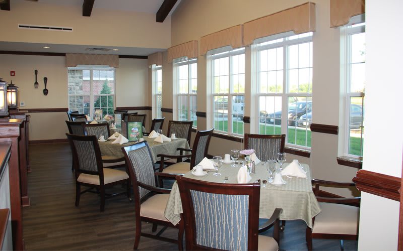 A well-lit dining room with neatly arranged tables and chairs, featuring large windows.