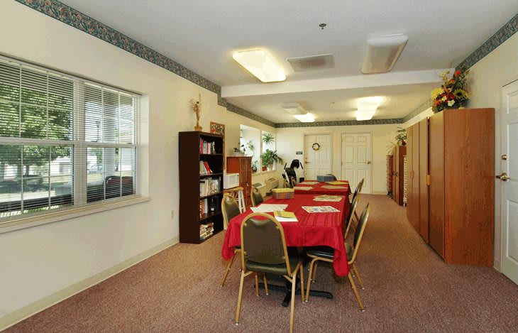 A well-lit meeting room with a conference table covered in a red cloth, surrounded by chairs and a bookshelf along the wall.