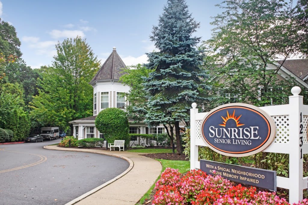 A picturesque building with surrounding trees and flowers, featuring a 'Sunrise Senior Living' sign by the pathway.
