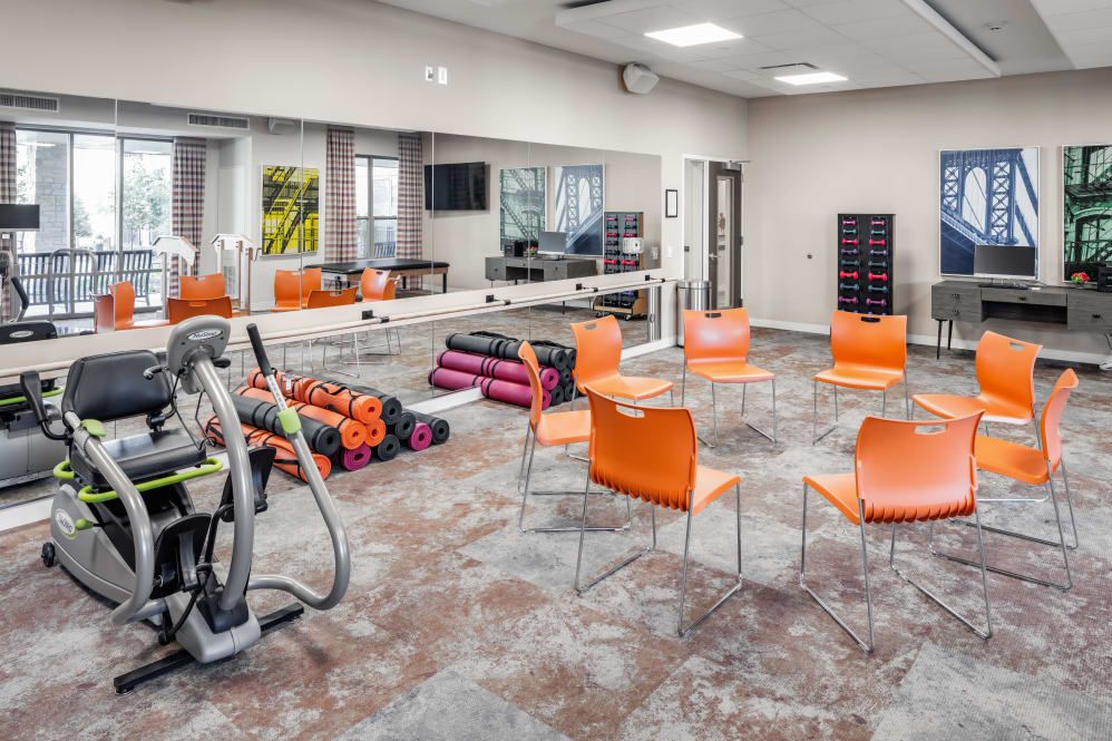 A brightly lit fitness room with exercise equipment, orange chairs, and mirrored walls.