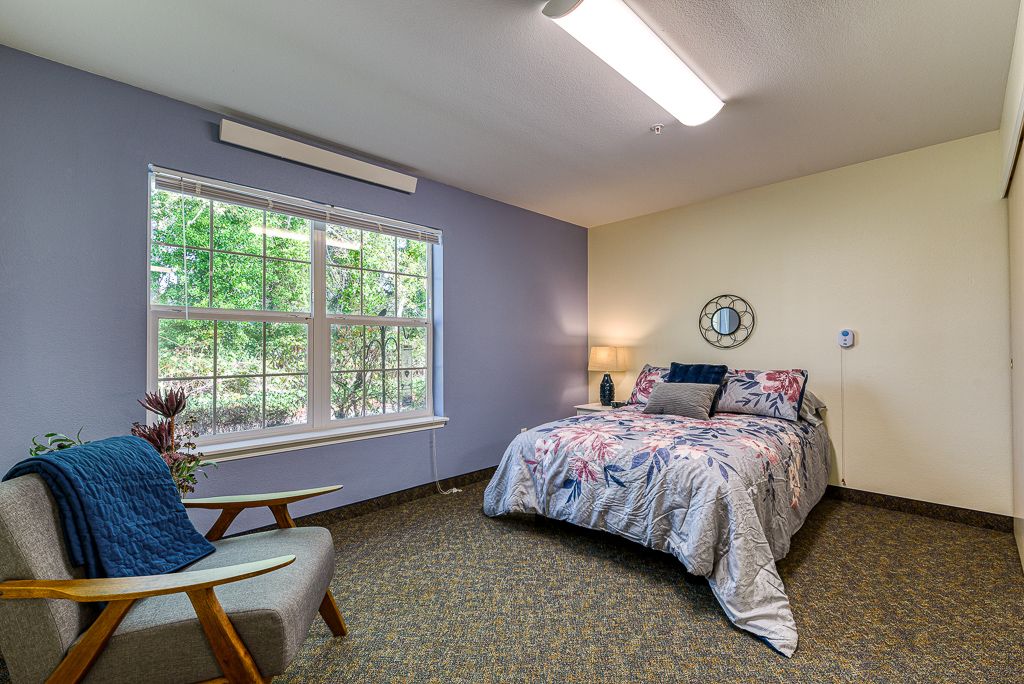 A cozy bedroom with a bed, chair, and large window letting in natural light.