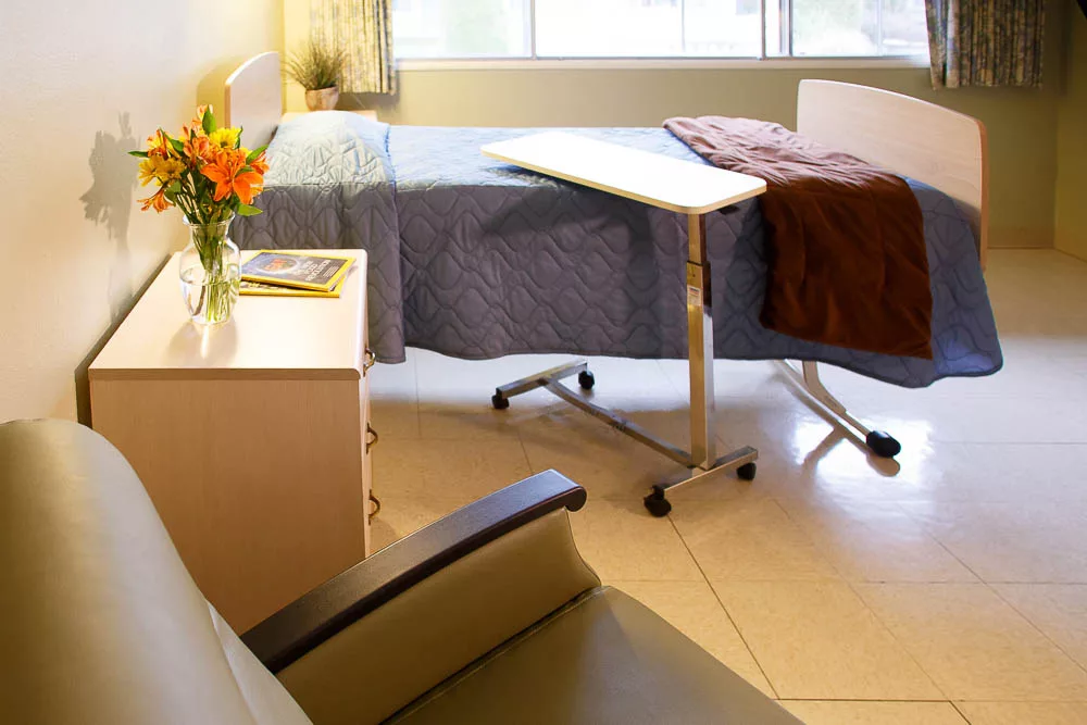 A well-lit hospital room with a bed, side table, and flowers by the window.