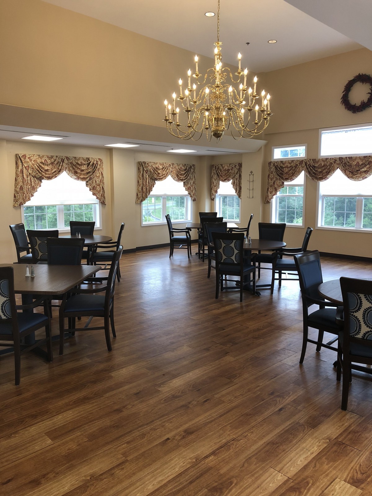 A dining area with several tables and chairs, featuring a chandelier and large windows with decorative curtains.