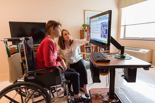 A healthcare professional assists a person in a wheelchair with technology at a desk.
