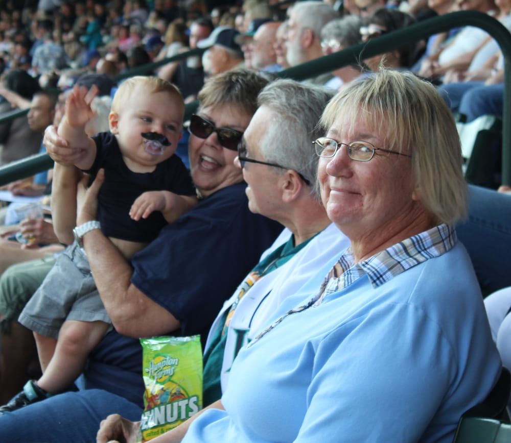 A baby with a fake mustache is held by smiling adults at a crowded stadium.