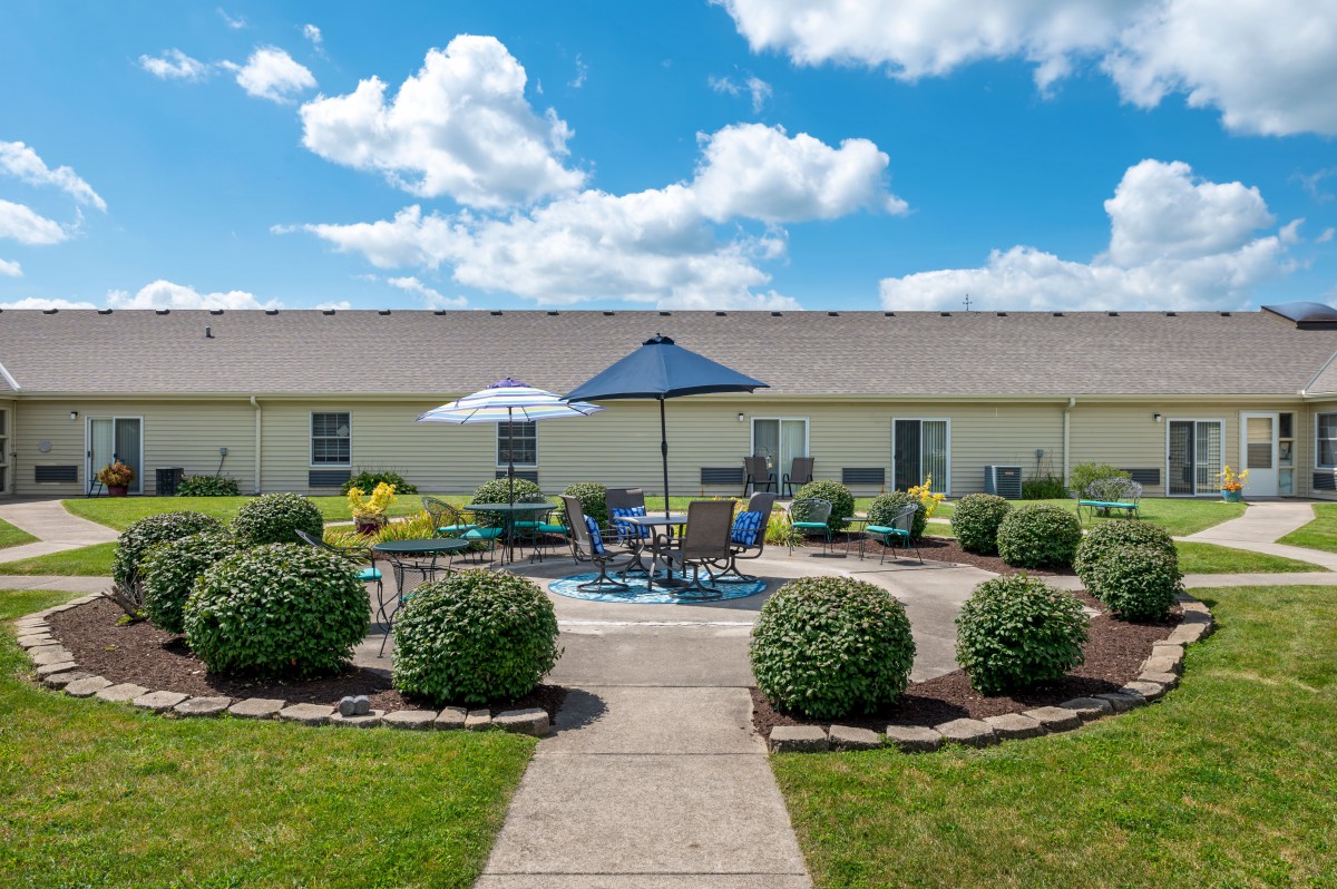 A well-maintained outdoor patio area with tables, chairs, and umbrellas surrounded by neatly trimmed bushes under a sunny blue sky.