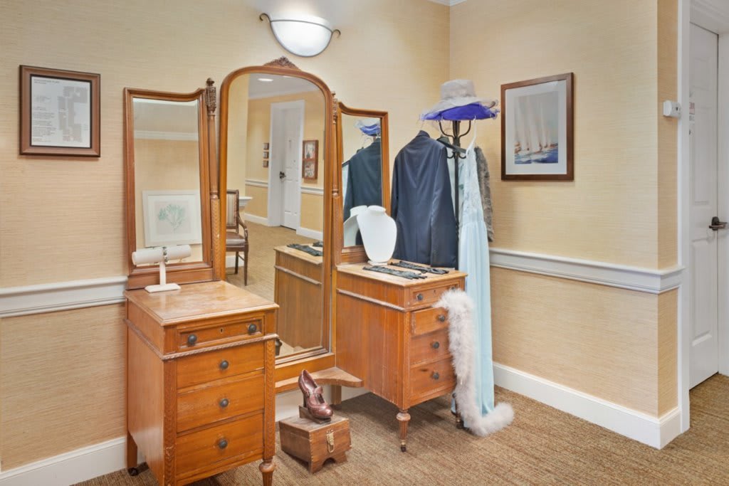 A vintage vanity desk with a mirror, accompanied by a coat rack holding various garments and accessories, creates an elegant room scene.