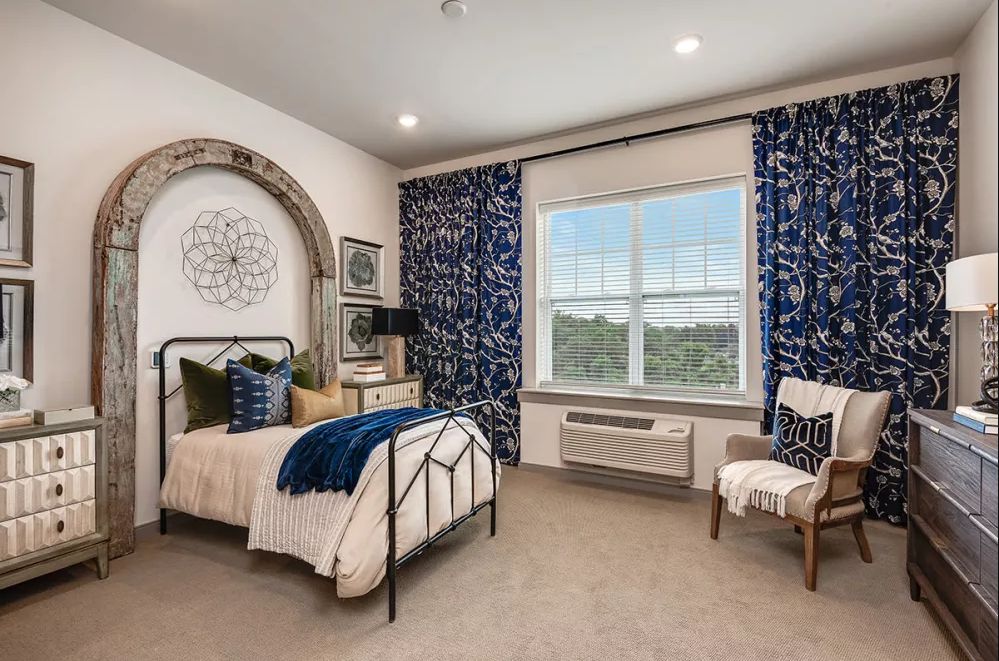 A well-decorated bedroom featuring a bed with blue and white accents, a large window with patterned curtains, and stylish furniture.