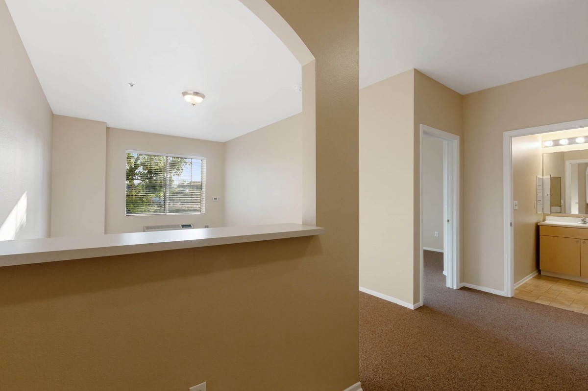 An empty apartment interior with beige walls, a carpeted floor, and an archway leading to another room.