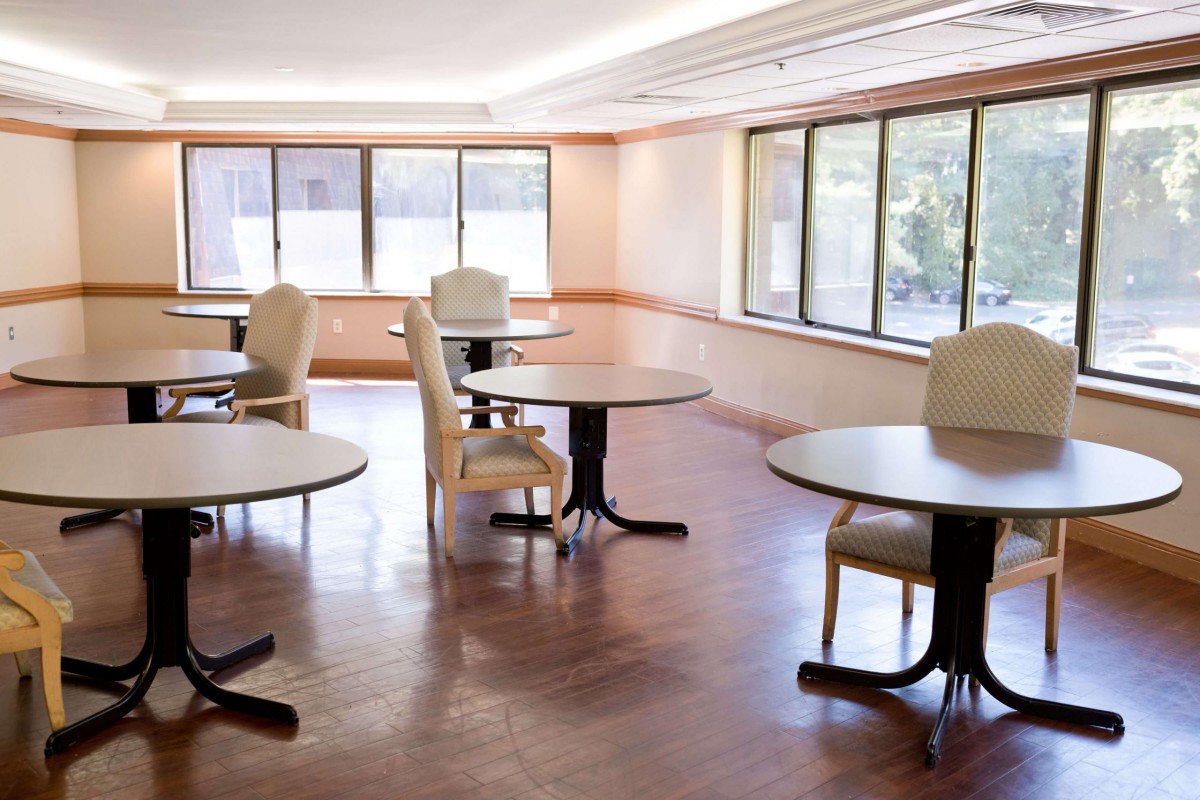 An empty room with tables and chairs, well-lit by natural light coming through large windows.