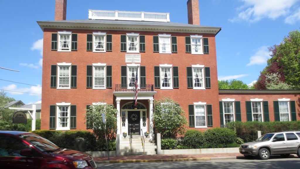 A historic brick building with green shutters and a central entrance, surrounded by plants.