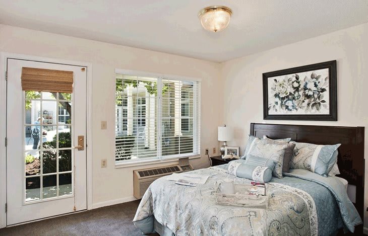 A cozy and well-lit bedroom with a neatly made bed, artwork on the wall, and a window offering natural light.