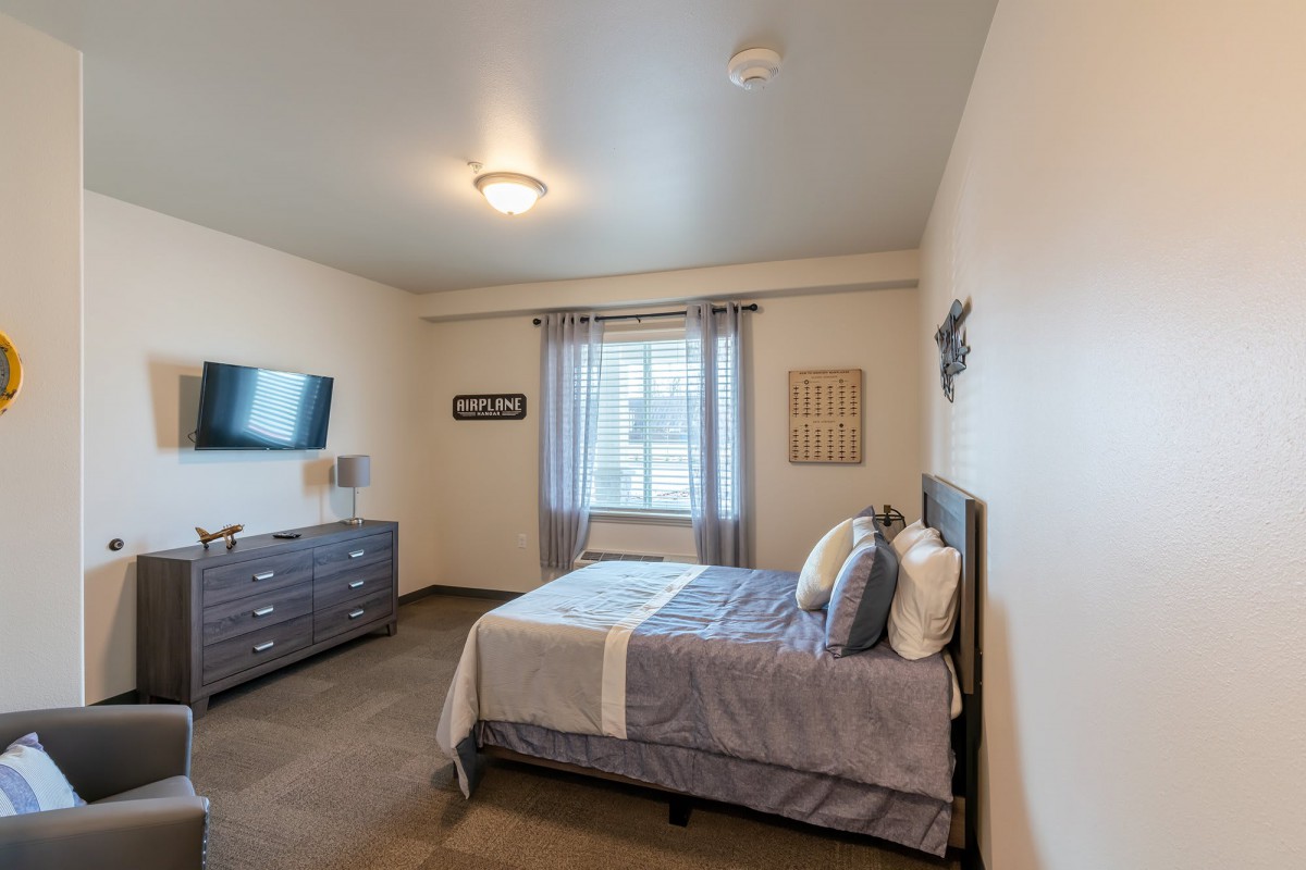 A modern bedroom featuring a neatly made bed, dresser with a television, and natural light streaming through a window with curtains.