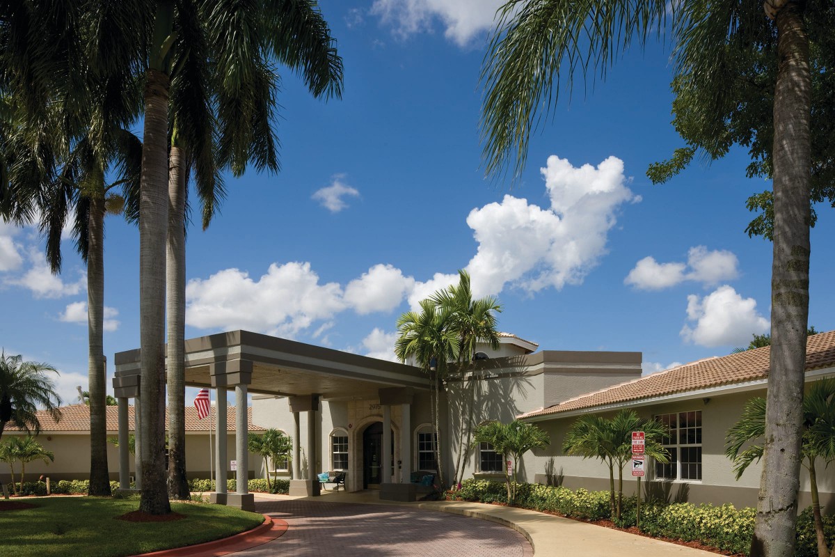 A building entrance surrounded by palm trees under a blue sky with clouds.