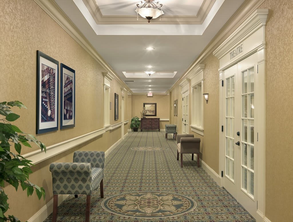 A well-lit hallway with elegant decor, featuring chairs, framed art and a patterned carpet.