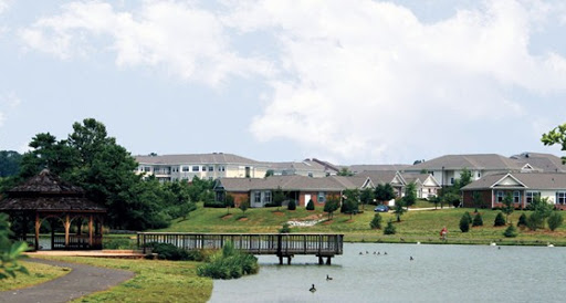 A serene view of an apartment complex by a lake, featuring a wooden bridge and lush greenery.