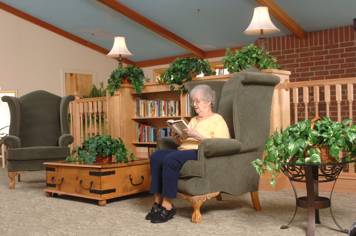An elderly woman sits comfortably in an armchair reading a book in a cozy library setting.