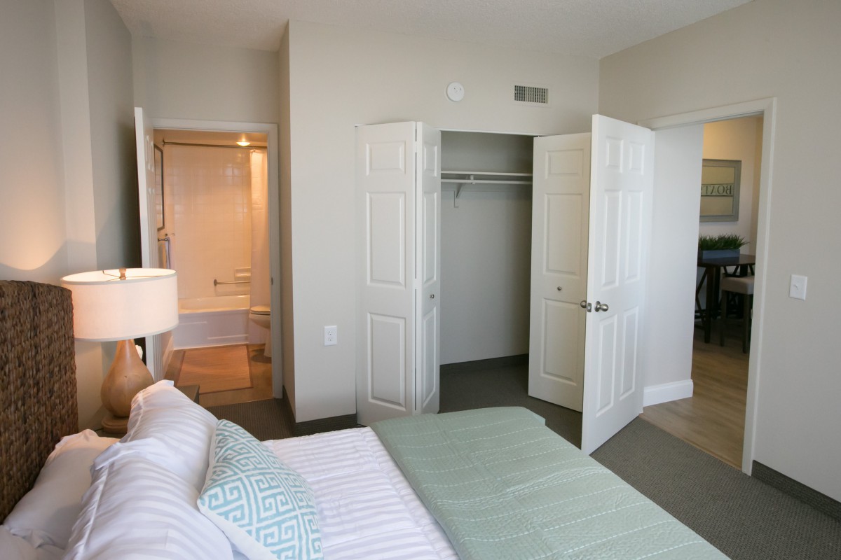 A modern, minimalist bedroom with a view into an adjacent bathroom; features include a neatly made bed and open closet doors.