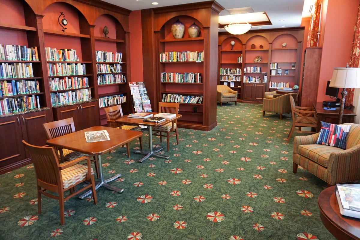 A cozy library with bookshelves, tables, and chairs set on a patterned carpet.