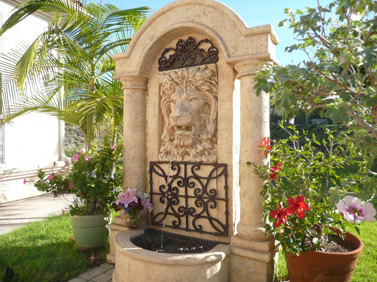 A decorative outdoor fountain with a lion sculpture, surrounded by plants in a sunny garden setting.