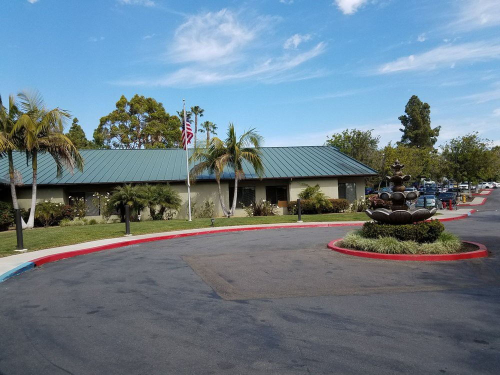 A building with a green roof surrounded by palm trees and landscaping, featuring a fountain in the driveway under a clear sky.