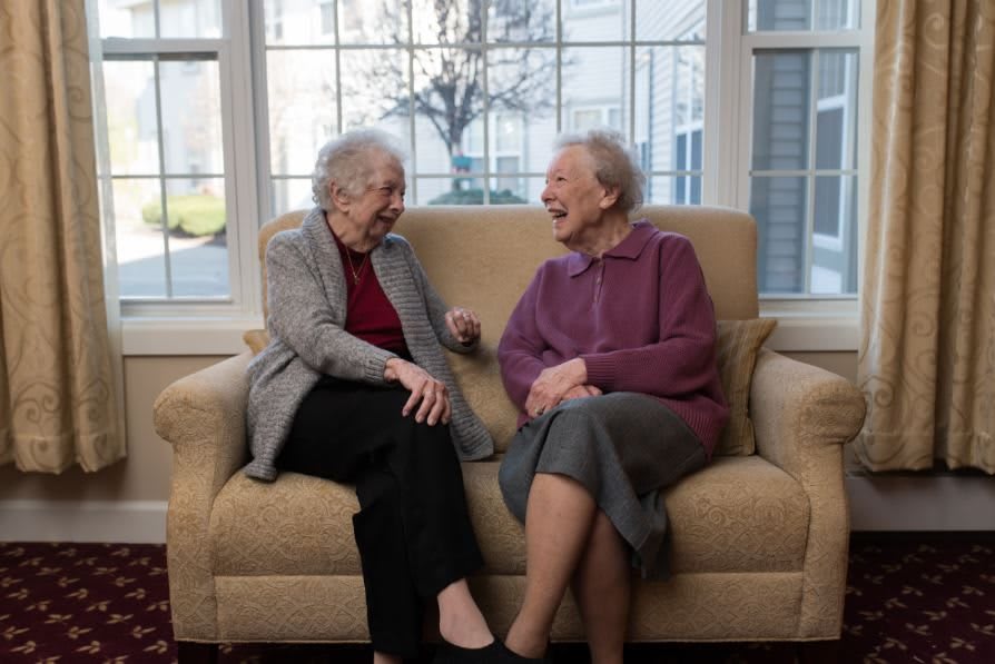 Two elderly women sitting on a couch, laughing and enjoying a conversation indoors.