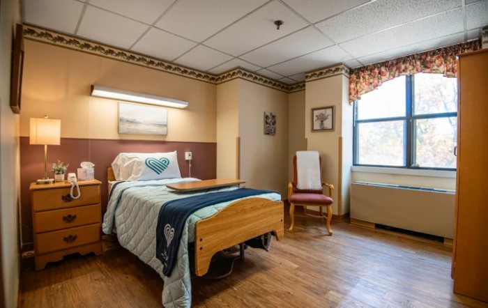 A cozy hospital room with a single bed and wooden furniture, featuring a window and a comfortable chair.