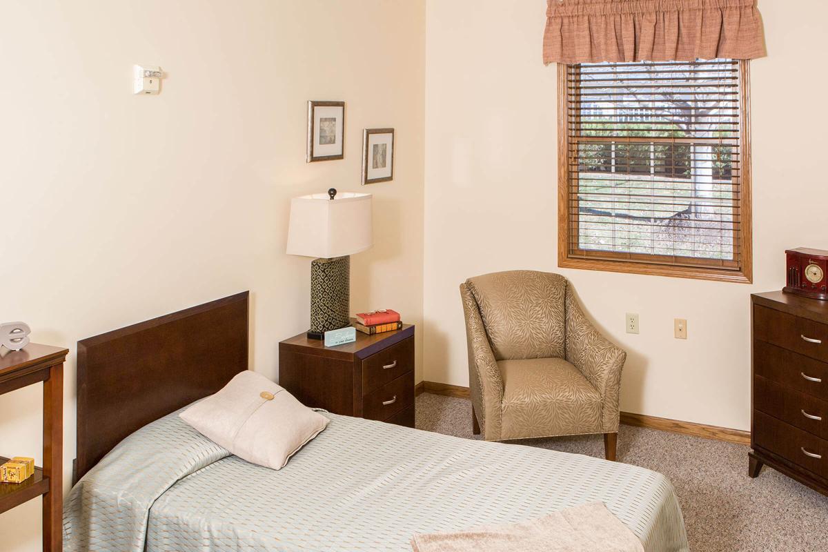 A simple bedroom with a single bed, a nightstand, and an armchair by the window.