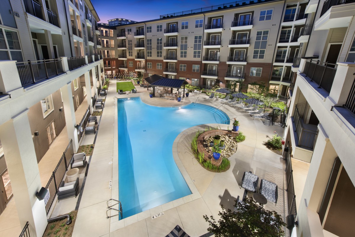 A modern apartment complex featuring a centrally located swimming pool and landscaped outdoor area, illuminated in the evening.